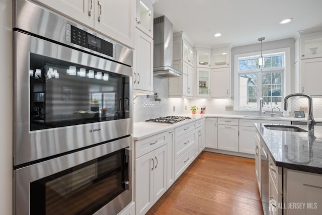 kitchen featuring light wood-type flooring, a sink, appliances with stainless steel finishes, white cabinets, and wall chimney range hood