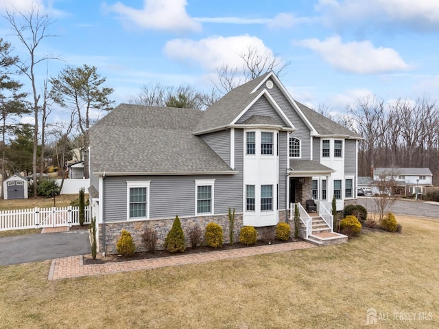 view of front of property featuring a front yard, fence, roof with shingles, stone siding, and aphalt driveway