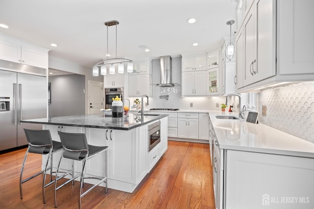 kitchen with a sink, light wood-style floors, built in appliances, white cabinetry, and wall chimney exhaust hood