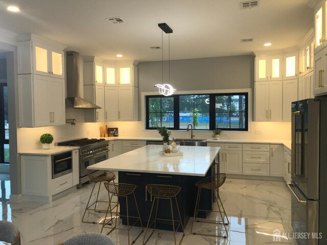 kitchen with appliances with stainless steel finishes, wall chimney exhaust hood, a kitchen island, decorative light fixtures, and white cabinetry