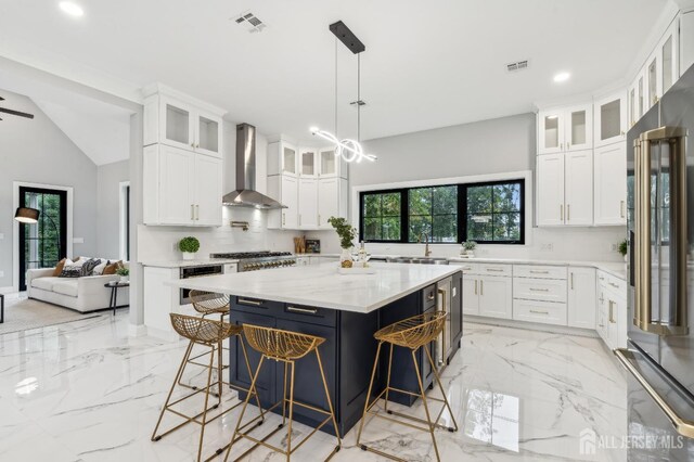 kitchen with wall chimney range hood, vaulted ceiling, appliances with stainless steel finishes, a kitchen island, and white cabinetry