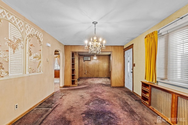 unfurnished dining area featuring arched walkways, a notable chandelier, dark carpet, wood walls, and baseboards