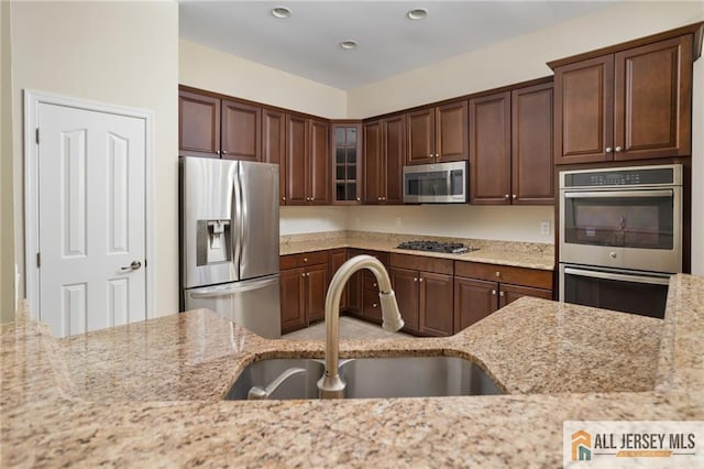 kitchen featuring a sink, dark brown cabinets, appliances with stainless steel finishes, light stone countertops, and glass insert cabinets