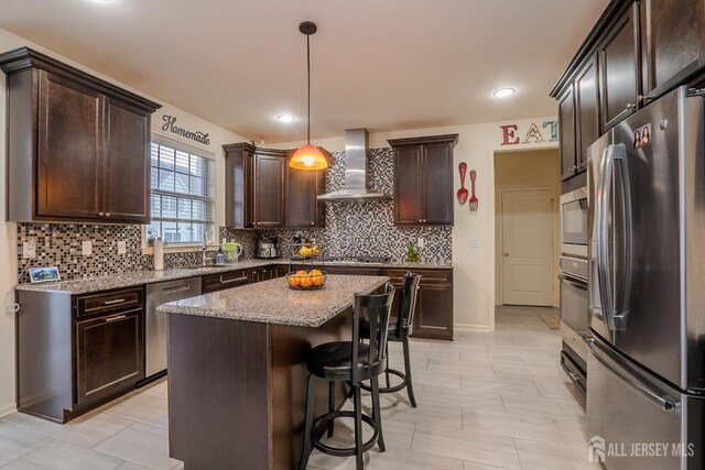 kitchen with appliances with stainless steel finishes, wall chimney exhaust hood, dark brown cabinetry, tasteful backsplash, and light stone counters