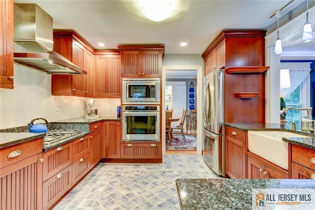 kitchen with wall chimney range hood, sink, hanging light fixtures, dark stone countertops, and stainless steel appliances