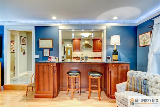 bar featuring ornamental molding, light wood-type flooring, dark stone counters, and wall chimney exhaust hood