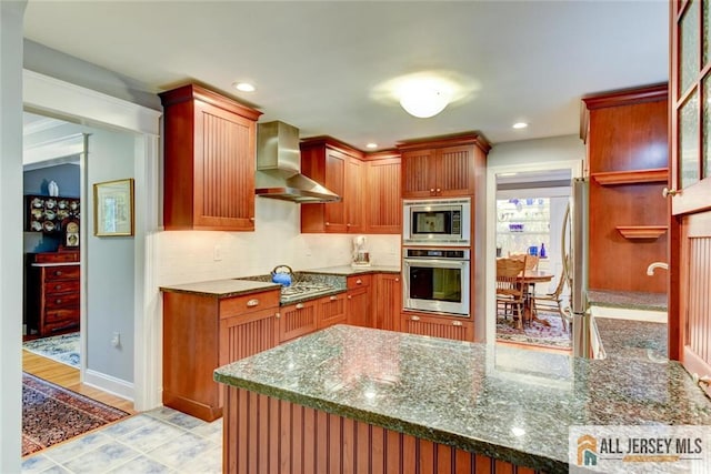 kitchen with wall chimney exhaust hood, light hardwood / wood-style flooring, kitchen peninsula, dark stone countertops, and appliances with stainless steel finishes