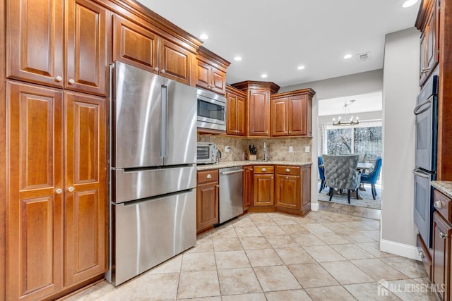 kitchen featuring stainless steel appliances, tasteful backsplash, light stone countertops, a chandelier, and brown cabinets