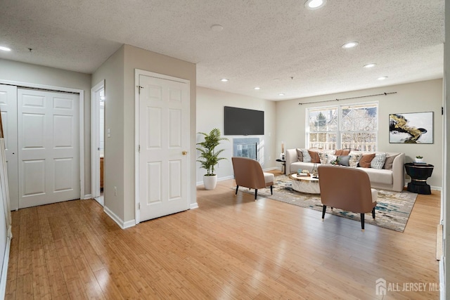 living area featuring light wood-type flooring, a textured ceiling, a glass covered fireplace, recessed lighting, and baseboards