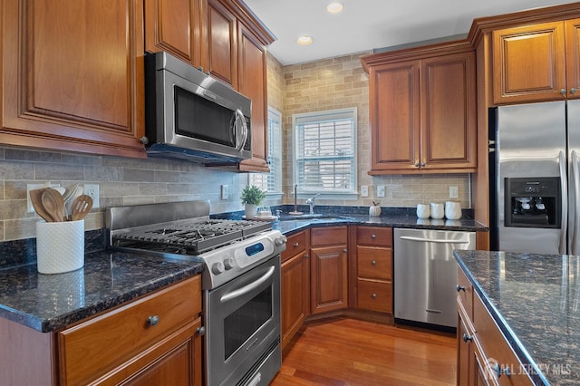 kitchen featuring light wood-style flooring, backsplash, stainless steel appliances, dark stone counters, and brown cabinetry