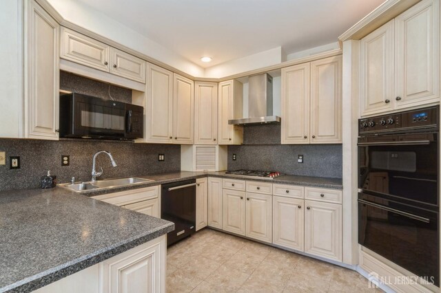 kitchen featuring dark countertops, wall chimney range hood, cream cabinets, black appliances, and a sink