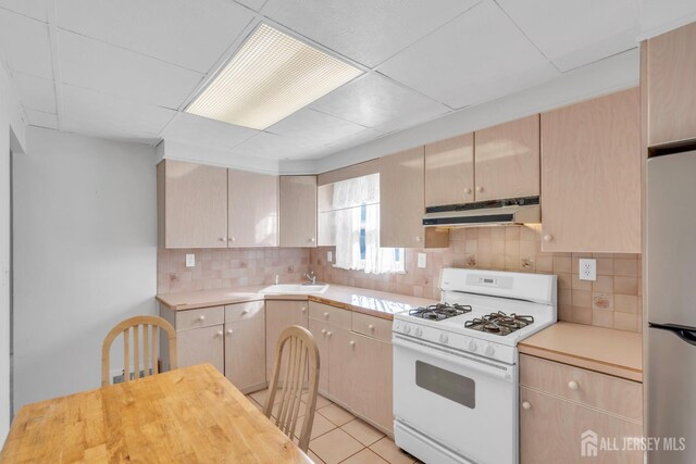 kitchen with stainless steel refrigerator, light brown cabinetry, white gas range oven, and light tile patterned floors