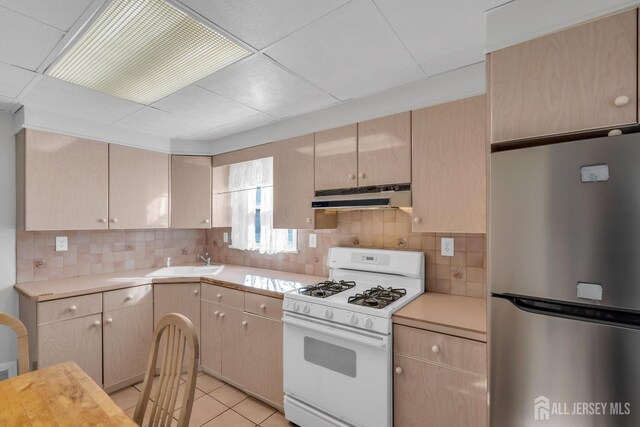 kitchen with sink, white gas stove, tasteful backsplash, light brown cabinetry, and stainless steel refrigerator