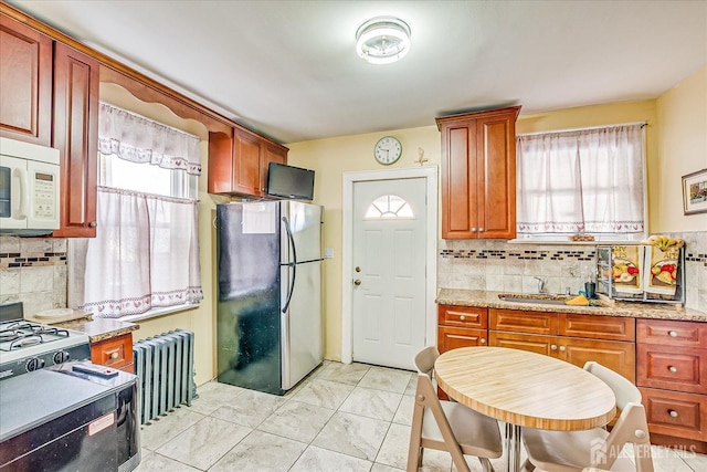 kitchen with radiator heating unit, sink, stainless steel fridge, decorative backsplash, and light stone countertops