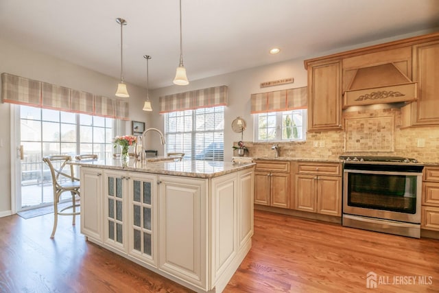 kitchen featuring decorative backsplash, custom exhaust hood, a kitchen island with sink, stainless steel range, and light hardwood / wood-style flooring