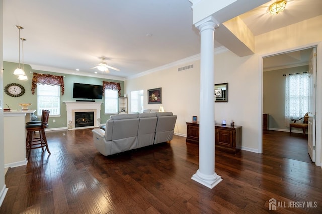 living room with ornate columns, a ceiling fan, crown molding, a glass covered fireplace, and dark wood-style floors