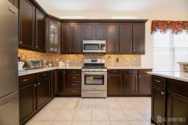 kitchen featuring stainless steel appliances, light countertops, backsplash, dark brown cabinets, and glass insert cabinets