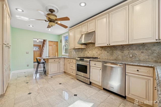kitchen with cream cabinetry, appliances with stainless steel finishes, under cabinet range hood, a peninsula, and baseboards