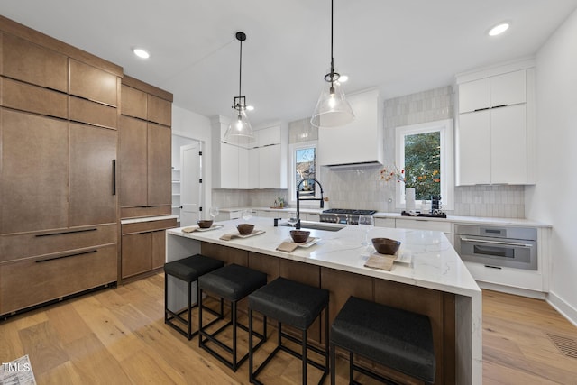 kitchen with sink, decorative light fixtures, an island with sink, oven, and white cabinets