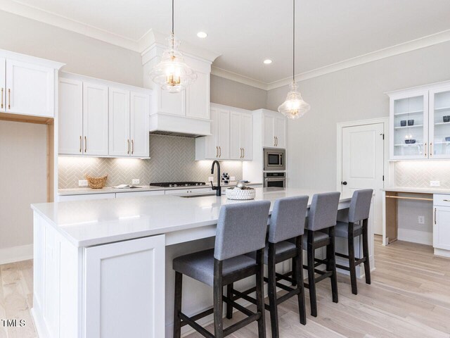 kitchen with glass insert cabinets, appliances with stainless steel finishes, a kitchen island with sink, light countertops, and pendant lighting