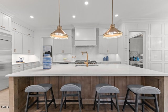 kitchen with stainless steel built in refrigerator, premium range hood, a large island with sink, pendant lighting, and white cabinets