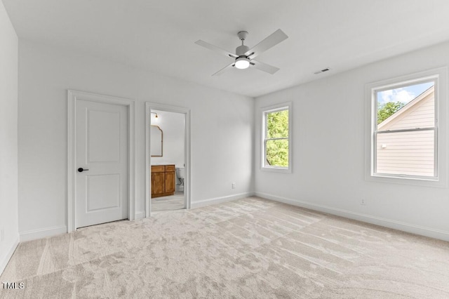 unfurnished bedroom featuring ensuite bathroom, multiple windows, light colored carpet, and ceiling fan