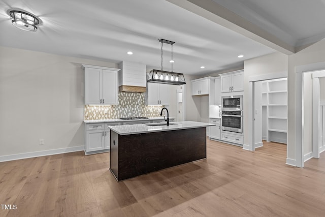 kitchen with sink, white cabinets, stainless steel appliances, custom range hood, and a center island with sink