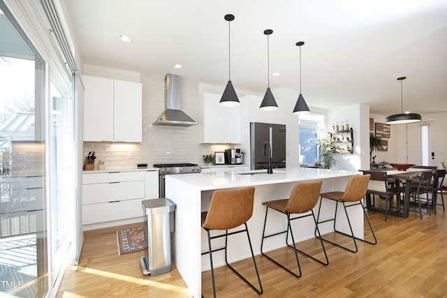 kitchen with pendant lighting, stainless steel appliances, a center island with sink, wall chimney exhaust hood, and white cabinets