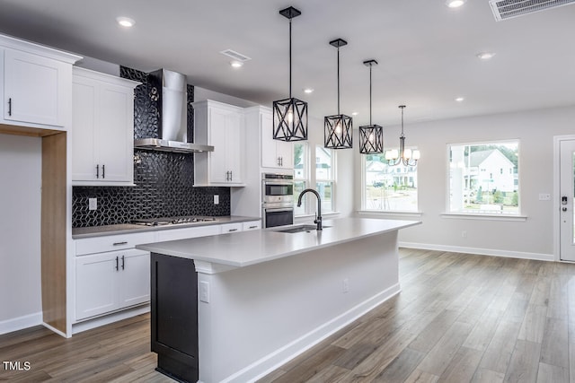 kitchen featuring decorative light fixtures, backsplash, wall chimney range hood, an island with sink, and hardwood / wood-style floors