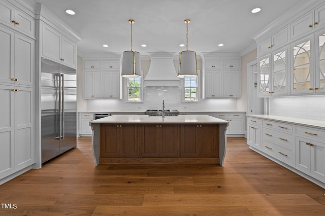 kitchen featuring wood-type flooring, premium range hood, white cabinetry, backsplash, and built in refrigerator