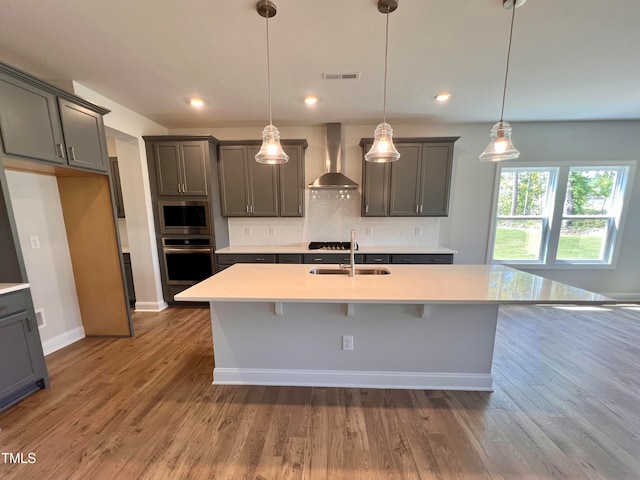 kitchen with sink, hanging light fixtures, wall chimney range hood, an island with sink, and appliances with stainless steel finishes