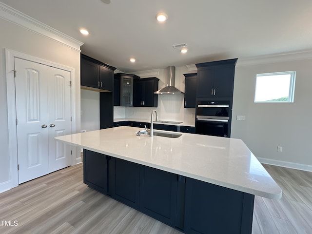 kitchen featuring wall chimney exhaust hood, double oven, an island with sink, sink, and light stone counters