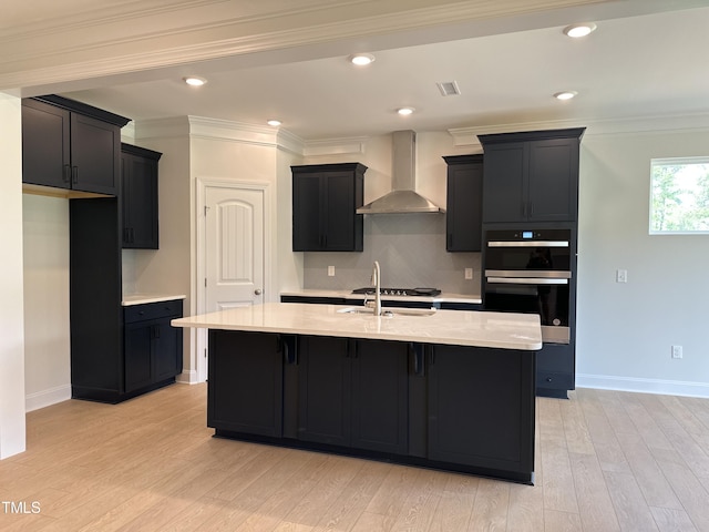 kitchen featuring an island with sink, wall chimney range hood, double oven, light hardwood / wood-style flooring, and sink