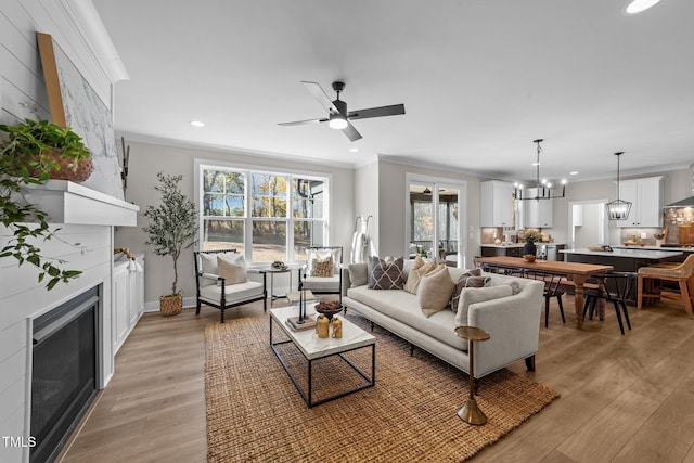 living room with crown molding, a fireplace, ceiling fan with notable chandelier, and light wood-type flooring