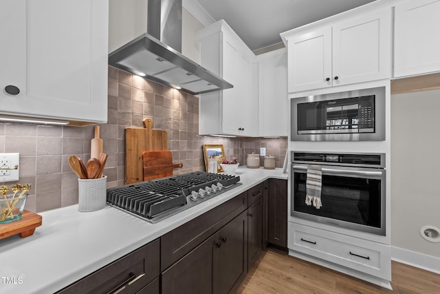 kitchen featuring dark brown cabinetry, white cabinetry, light wood-type flooring, stainless steel appliances, and wall chimney range hood