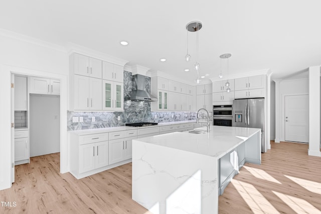 kitchen with sink, wall chimney exhaust hood, hanging light fixtures, an island with sink, and white cabinets