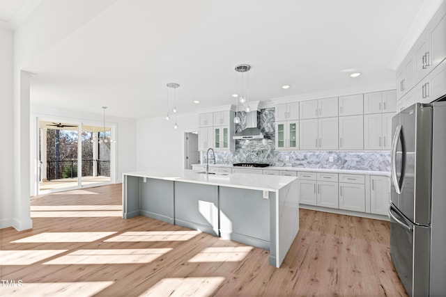 kitchen with stainless steel fridge, a center island with sink, hanging light fixtures, and wall chimney range hood