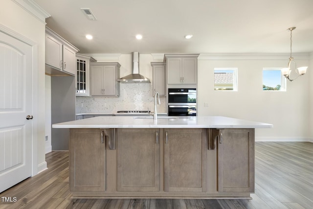 kitchen with wall chimney exhaust hood, wood-type flooring, sink, and a large island with sink