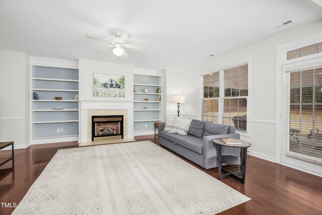 living room featuring visible vents, built in shelves, a fireplace with raised hearth, baseboards, and dark wood-style flooring