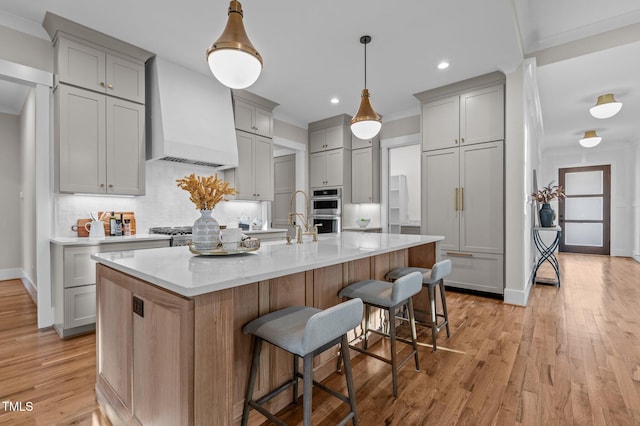 kitchen featuring pendant lighting, a center island with sink, stainless steel double oven, premium range hood, and a kitchen breakfast bar