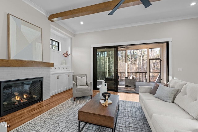 living room featuring a ceiling fan, crown molding, light wood-style floors, a fireplace, and beam ceiling