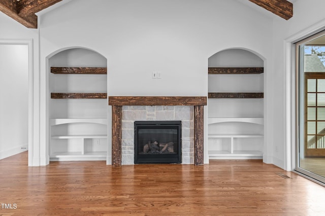 unfurnished living room with built in shelves, a stone fireplace, lofted ceiling with beams, and hardwood / wood-style floors