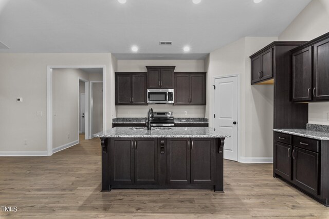 kitchen with appliances with stainless steel finishes, light hardwood / wood-style flooring, a kitchen island with sink, and light stone counters