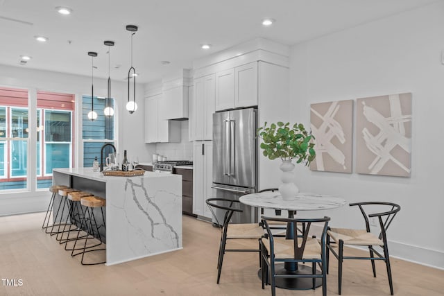 kitchen with light stone counters, stainless steel appliances, white cabinetry, an island with sink, and custom range hood