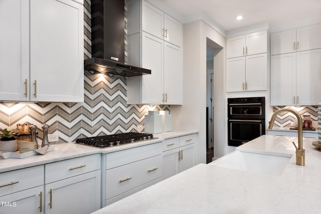 kitchen featuring light stone countertops, white cabinets, black double oven, wall chimney range hood, and sink