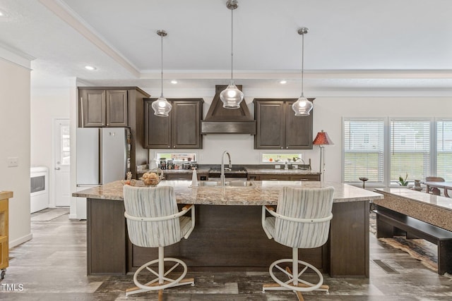 kitchen with custom range hood, light stone countertops, a breakfast bar, and an island with sink