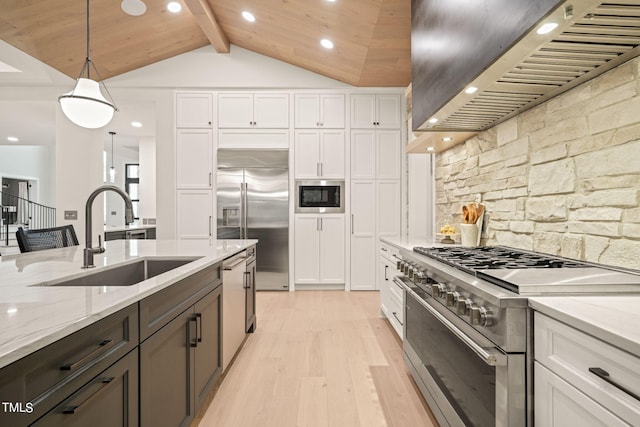 kitchen featuring built in appliances, a sink, white cabinets, wall chimney range hood, and decorative light fixtures