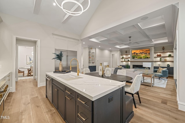 kitchen featuring ceiling fan, an island with sink, sink, coffered ceiling, and light wood-type flooring