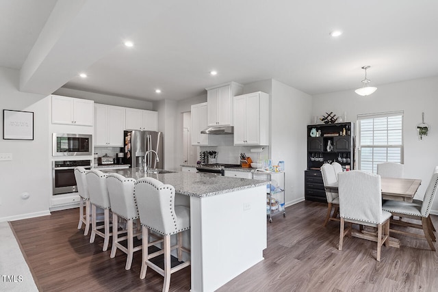 kitchen with stainless steel appliances, wood-type flooring, a center island with sink, light stone countertops, and white cabinetry