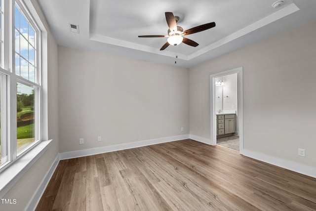 unfurnished bedroom featuring ceiling fan, ensuite bath, a tray ceiling, and light hardwood / wood-style flooring